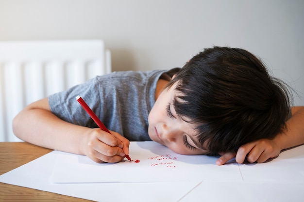 Portret van kind dat een brief schrijft, Weinig jongen die rood hart op Witboek trekt voor zijn mamma.