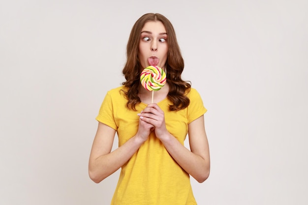 Portret van kinderachtig tienermeisje met krullend haar in stedelijke stijl T-shirt likken lolly met gekruiste ogen, genieten van zoet, heeft een grappige uitdrukking. Indoor studio opname geïsoleerd op een grijze achtergrond.