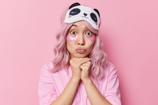 Portret van knappe Aziatische vrouw houdt handen onder kin lippen gevouwen past schoonheidspleisters toe voor huidbehandeling draagt slaapmasker op voorhoofd en casual t-shirt geïsoleerd over roze achtergrond