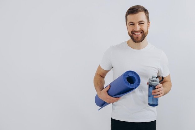 Portret van knappe bebaarde man in sportkleding met yoga mat en fles vers water op witte achtergrond kopie ruimte