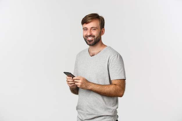 Portret van knappe bebaarde man met een smartphone, glimlachend tevreden op camera, permanent over wit
