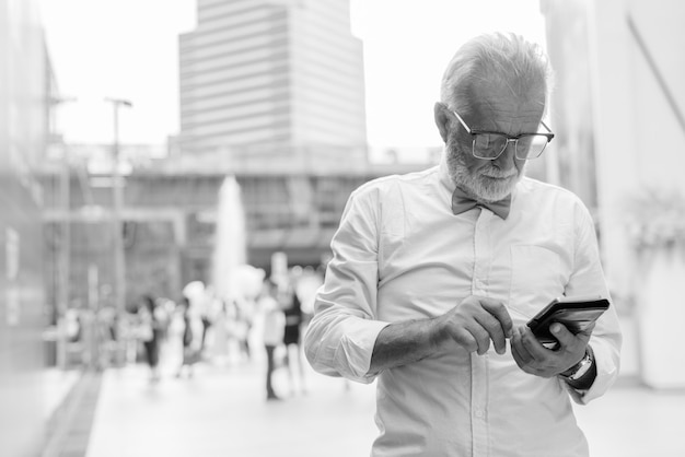 Portret van knappe bebaarde senior toeristische man stijlvolle kleding dragen tijdens het verkennen van de stad Bangkok in zwart-wit