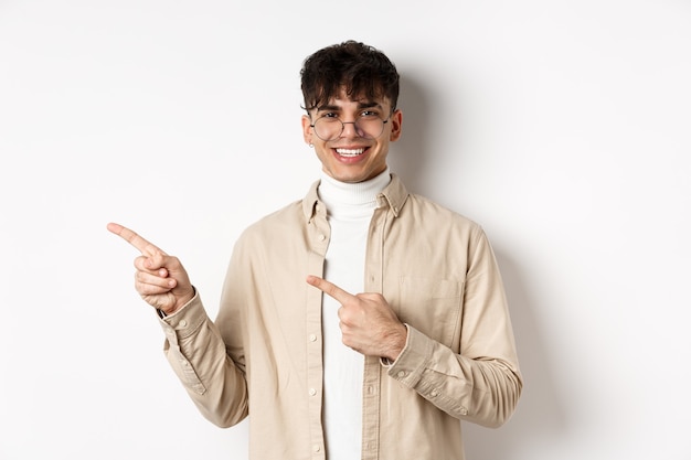Portret van knappe blanke jongeman in bril met logo, glimlachend en wijzende vingers links, uitnodigend om promo deal, witte muur te bekijken.