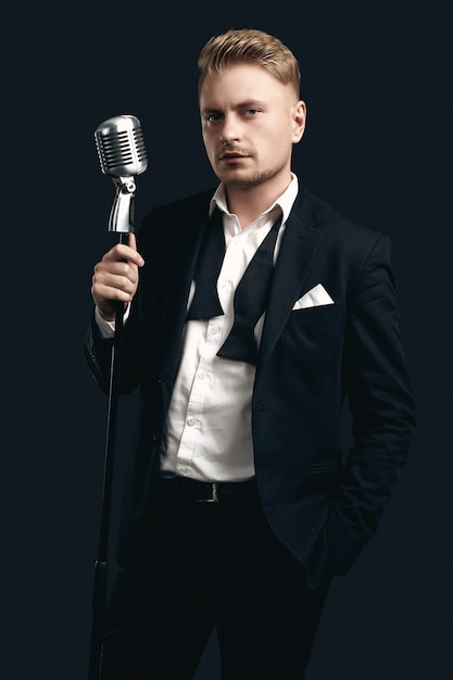 Foto portret van knappe blonde man zanger in elegante smoking en vlinderdas poseren met vintage microfoon op zwarte muur