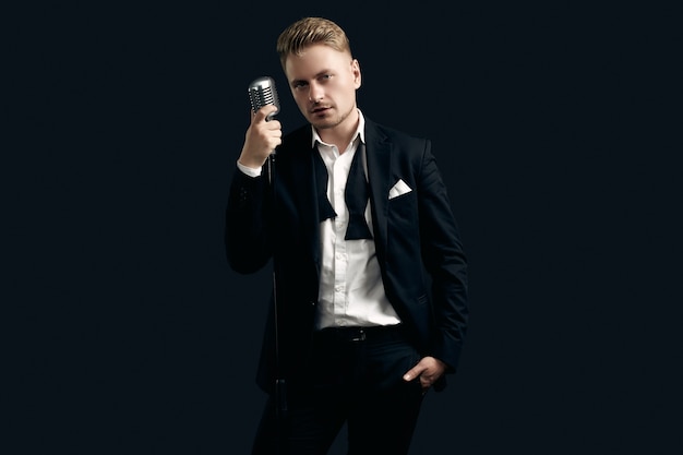 Foto portret van knappe blonde man zanger in elegante smoking en vlinderdas poseren met vintage microfoon op zwarte muur