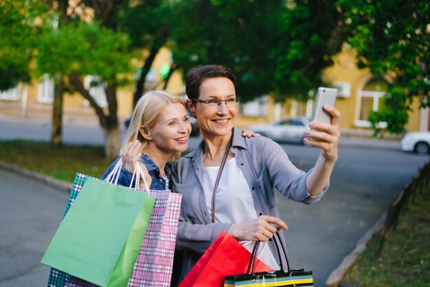 Portret van knappe dames die buiten selfies maken met boodschappentassen