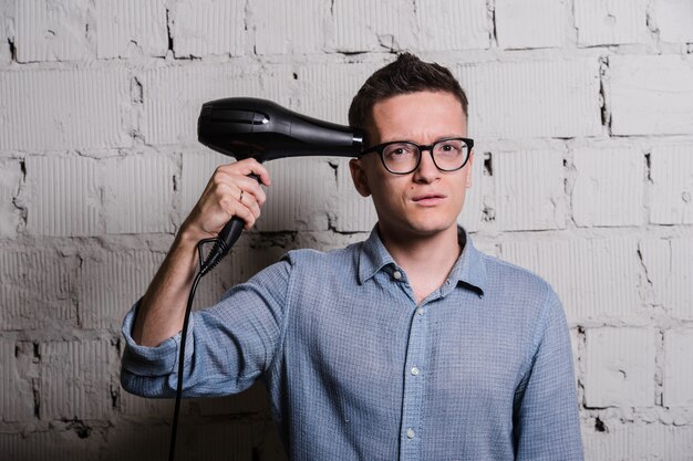 Portret van knappe jonge man in jeans kleding en bril met haardroger op zoek naar camera glimlachen, staande tegen de grijze bakstenen muur