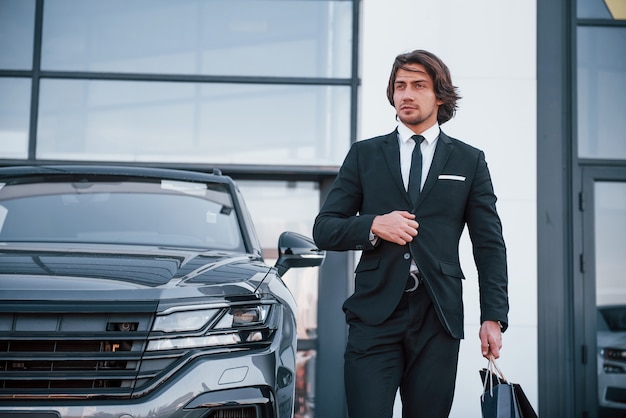 Portret van knappe jonge zakenman in zwart pak en stropdas buiten in de buurt van moderne auto en met boodschappentassen.