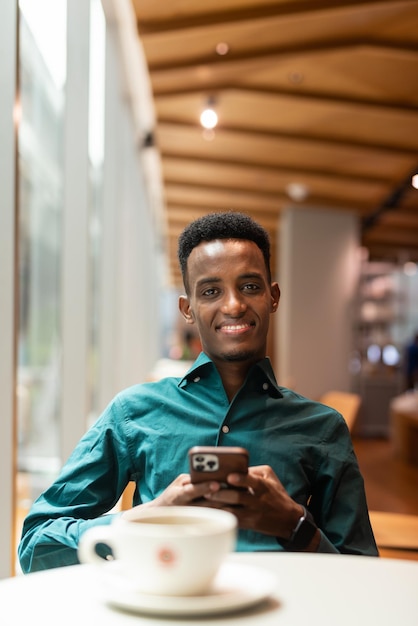 Portret van knappe jonge zwarte man in coffeeshop met behulp van telefoon