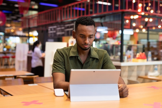 Portret van knappe jonge zwarte man met behulp van laptopcomputer in coffeeshop