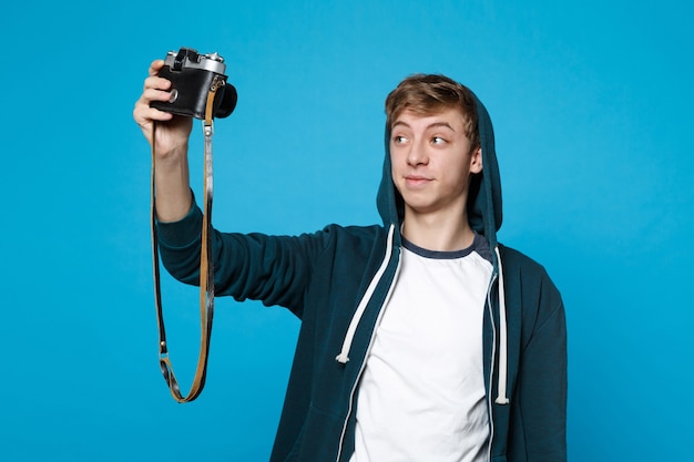 Portret van knappe jongeman in casual kleding die selfie geschoten op retro vintage fotocamera geïsoleerd op blauwe muur doet. Mensen oprechte emoties, lifestyle concept.
