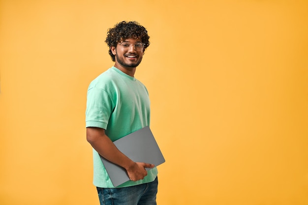 Portret van knappe krullende Indiase man in turquoise t-shirt en bril lachend naar camera kijkend en laptop vasthoudend