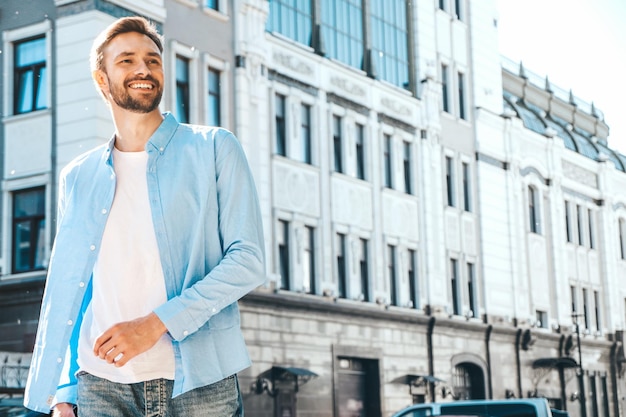 Portret van knappe lachende stijlvolle hipster lamberseksueel model moderne man gekleed in blauw shirt mode man poseren in de buurt van wolkenkrabber op de straat achtergrond buitenshuis bij zonsondergang