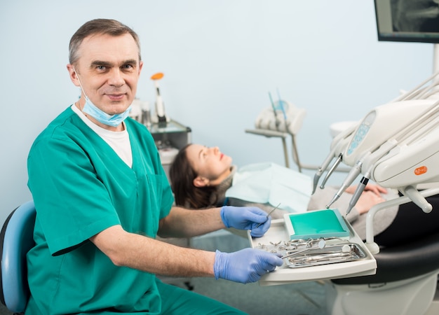 Portret van knappe mannelijke tandarts met tandheelkundige apparaten in de tandheelkundige kliniek. Vrouwelijke patiënt. Tandheelkunde