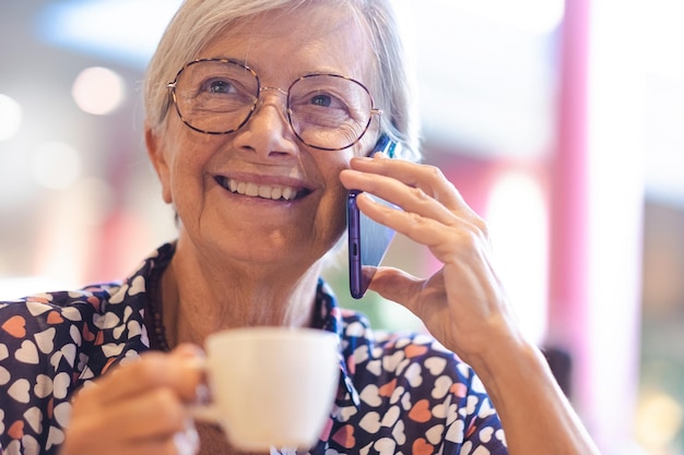 Portret van knappe senior vrouw in blauw shirt praten op mobiele telefoon tijdens het drinken van koffie in cafetaria Glimlachende vrouw met behulp van slimme telefoon terwijl het drinken van espresso