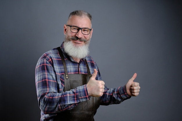 Portret van knappe volwassen bebaarde man gekleed lederen schort gebaren duimen omhoog over grijze muur Kaukasische werkman met baard glimlachen