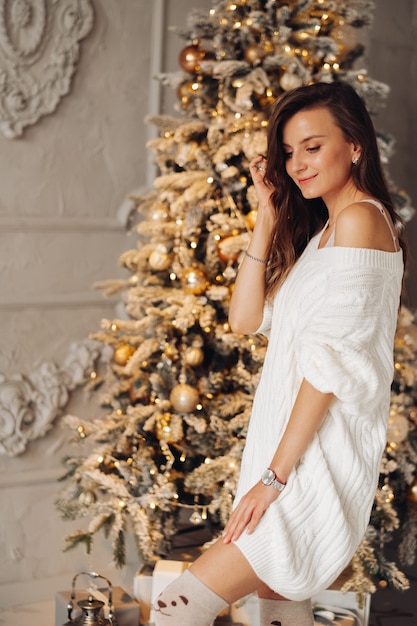 Portret van knappe vrouw in witte jurk glimlacht in de buurt van de kerstboom