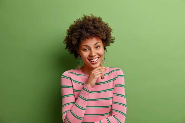 Portret van knappe vrouw met krullend haar houdt hand onder de kin glimlacht in het algemeen heeft tevreden gezichtsuitdrukking gekleed in casual gestreepte trui geïsoleerd over groene muuremotions