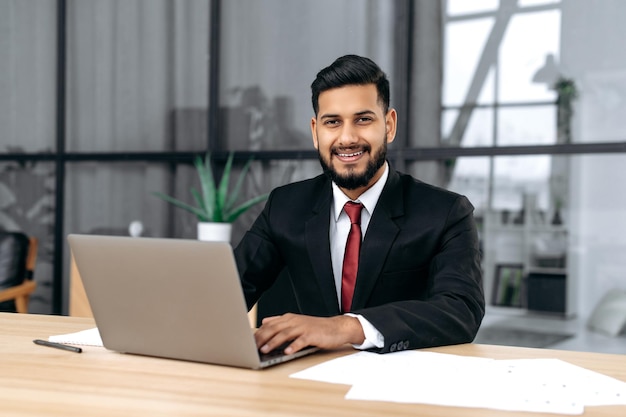 Portret van knappe zelfverzekerde Arabische of Indiase succesvolle zakenman ondernemer advocaat in een formeel pak zit aan het bureau met laptop in modern creatief kantoor kijkt naar camera glimlach vriendelijk