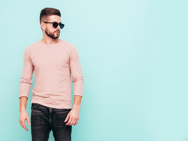 Portret van knappe zelfverzekerde model Sexy stijlvolle man gekleed in roze trui en jeans Fashion hipster man poseren in de buurt van blauwe muur in studio In zonnebril geïsoleerd