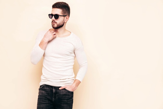 Portret van knappe zelfverzekerde stijlvolle hipster lamberseksueel model Man gekleed in witte trui en jeans Mode man poseren in de buurt van muur in studio in zonnebril