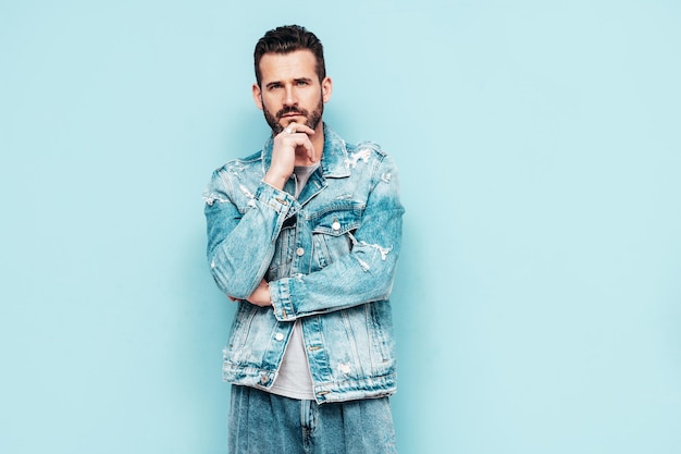 Portret van knappe zelfverzekerde stijlvolle hipster lamberseksueel model Sexy man gekleed in jeans jasje Mode man geïsoleerd op blauwe muur in studio