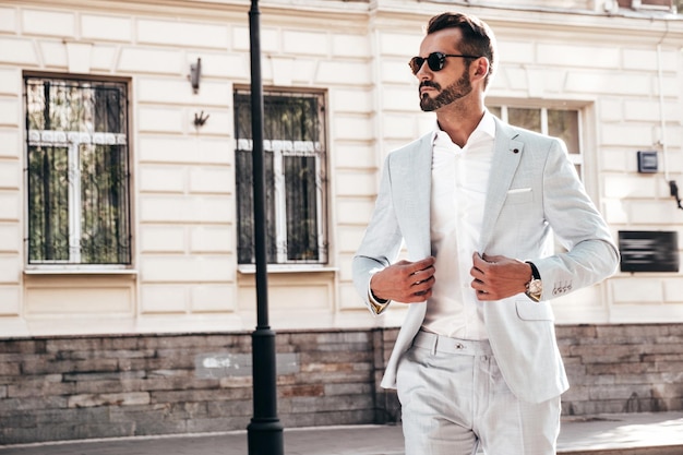 Portret van knappe zelfverzekerde stijlvolle hipster lamberseksueel modelSexy moderne man gekleed in elegant wit pak Fashion man poseren op de straat achtergrond in Europa stad bij zonsondergang In zonnebril