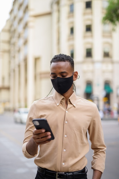 Portret van knappe zwarte jonge Afrikaanse zakenman die vrijetijdskleding buiten in stad draagt en mobiele telefoon gebruikt