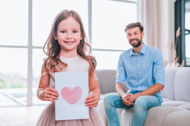 Portret van lachend meisje met een foto van het hart terwijl vader thuis op de bank zit