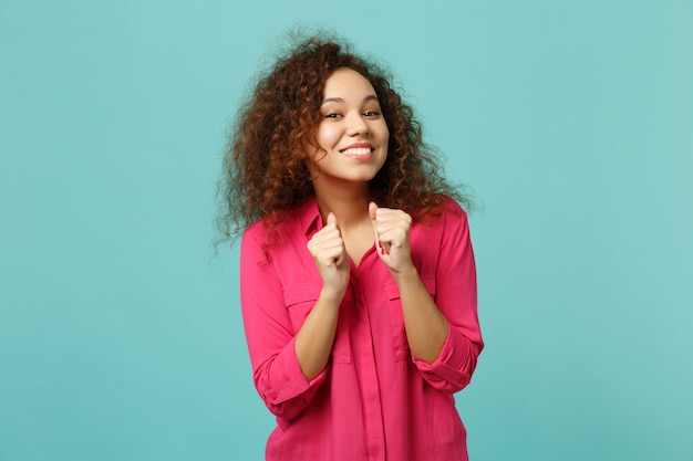 Portret van lachend vrij Afrikaans meisje in roze casual kleding balde vuisten geïsoleerd op blauwe turquoise muur achtergrond in studio. Mensen oprechte emoties, lifestyle concept. Bespotten kopie ruimte.