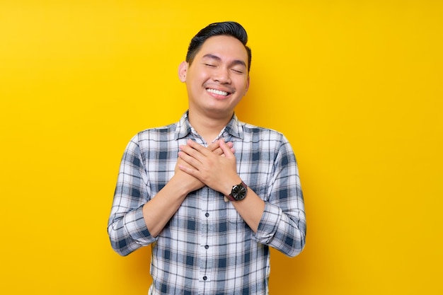 Portret van lachende aangename jonge aziatische man in geruite hemd camera kijken en hand in hand op zijn borst hart geïsoleerd op gele achtergrond mensen levensstijl concept