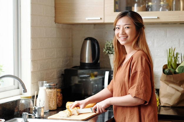 Portret van lachende aantrekkelijke jonge vrouw die brood snijdt voor het ontbijt