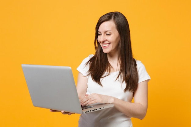 Portret van lachende aantrekkelijke jonge vrouw in witte casual kleding met behulp van laptop pc-computer geïsoleerd op fel geel oranje muur achtergrond in studio. Mensen levensstijl concept. Bespotten kopie ruimte.