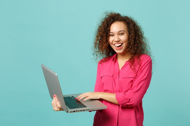 Portret van lachende Afrikaanse meisje in roze casual kleding met behulp van laptop pc-computer geïsoleerd op blauwe turquoise muur achtergrond in studio. Mensen oprechte emoties, lifestyle concept. Bespotten kopie ruimte.