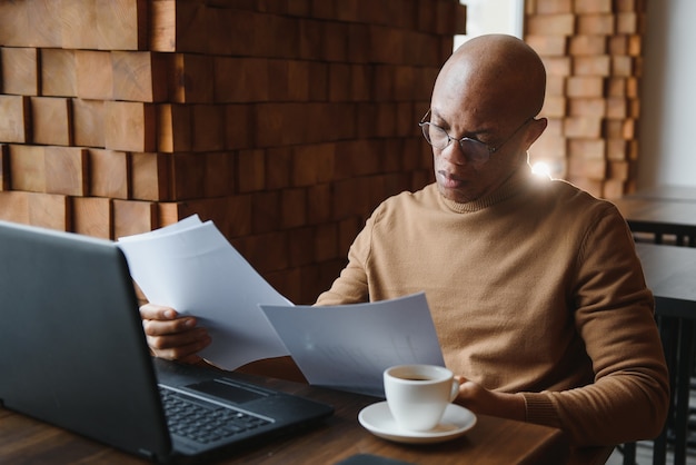 Portret van lachende Afro-Amerikaanse man met bril zit aan bureau in kantoor werken op laptop, gelukkige biraciale mannelijke werknemer kijkt naar camera poseren, druk bezig met het gebruik van moderne computergadget op werkplek