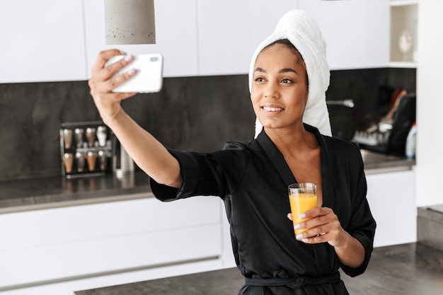 Portret van lachende Afro-Amerikaanse vrouw, gekleed in housecoat, selfie te nemen op de mobiele telefoon in de keuken