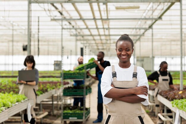 Portret van lachende Afro-Amerikaanse werknemer in kas met hydrocultuur omgeving met landbouwingenieurs met behulp van laptopcomputer. Vrouw poseren gelukkig in biologische gewassen en groenten boerderij.