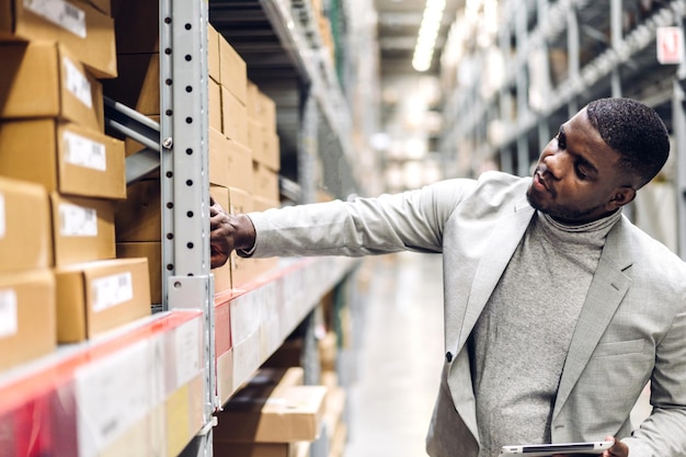 Portret van lachende Afro-Amerikaanse zakenman besteldetails op tablet die goederen en benodigdheden op planken controleert met goederenachtergrond in magazijnlogistiek en zakelijke export