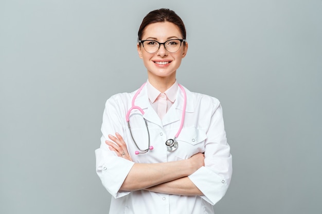 Foto portret van lachende arts in wit uniform staande met gekruiste handen op grijze achtergrond