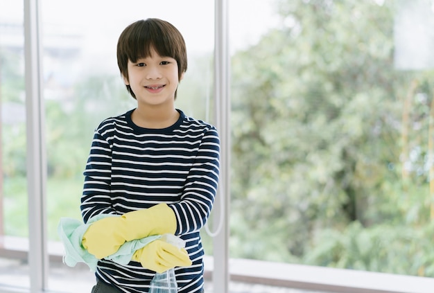 Portret van lachende Aziatische jongen met spuitfles en vod die rubberen handschoenen draagt en camera thuis bekijkt Jongen helpt bij het schoonmaken van het huis