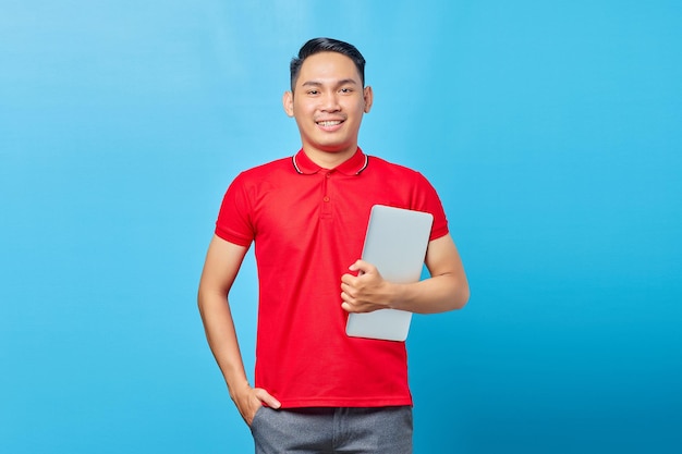 Portret van lachende Aziatische knappe jonge man in rood shirt met laptop en kijken naar camera geïsoleerd op blauwe achtergrond