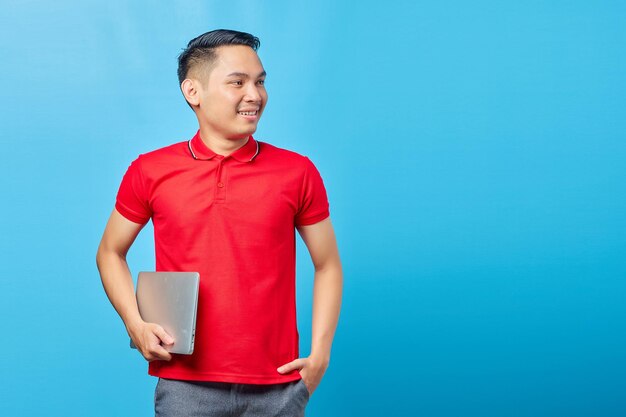 Portret van lachende Aziatische knappe jonge man in rood shirt met laptop en kijken naar kopieerruimte geïsoleerd op blauwe achtergrond