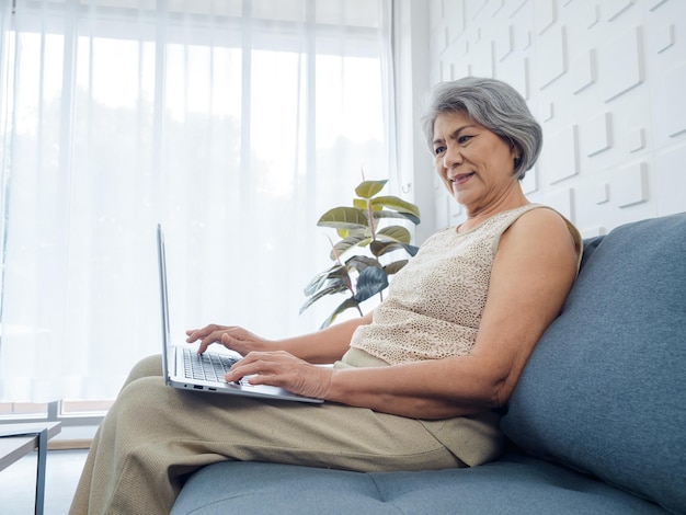 Portret van lachende aziatische senior casual vrouw zittend op de bank werken met laptopcomputer in helder witte kamer oudere vrouw surfen op internet of videogesprek thuis ouderen met technologie