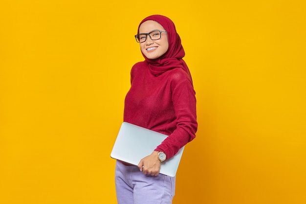 Portret van lachende Aziatische vrouw brengt laptop over gele achtergrond