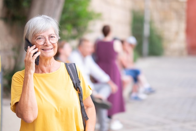 Portret van lachende blanke senior vrouw in gele t-shirt en bril die buiten staat te praten op mobiele telefoon