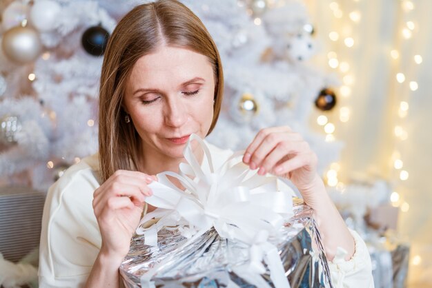 Portret van lachende blanke vrouw met zilveren geschenkdoos in de buurt van boom een jonge vrouw opent haar cadeau...