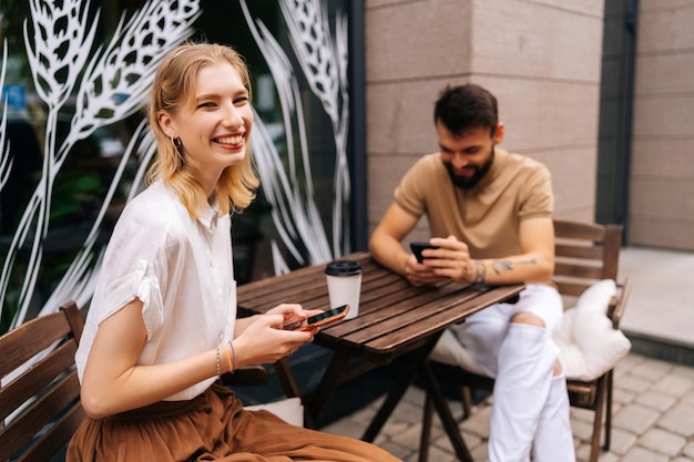 Portret van lachende blonde jonge vrouw kijkend naar camera bebaarde getatoeëerde man met behulp van mobiele telefoon kijkend naar scherm zittend aan tafel met koffie