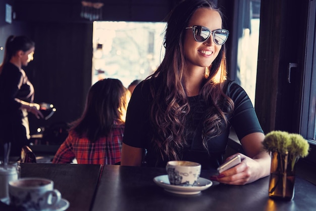 Portret van lachende brunette vrouw in zonnebril, drinkt koffie in de ochtend in een café.