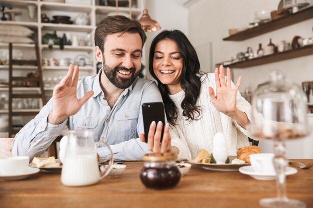 Portret van lachende europese paar man en vrouw die smartphone gebruiken terwijl ze thuis ontbijten in de keuken