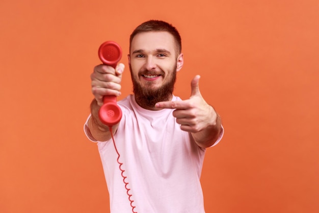 Portret van lachende gelukkig bebaarde man wijzende vinger op retro telefoon handset naar camera vragen om de telefoon te beantwoorden dragen roze tshirt indoor studio shot geïsoleerd op oranje achtergrond